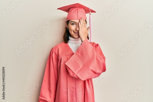 Young caucasian woman wearing graduation cap and ceremony robe covering one eye with hand, confident smile on face and surprise emotion.