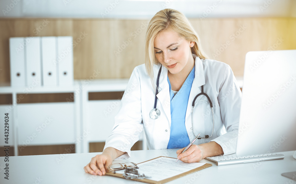 Woman-doctor at work while sitting at the desk in sunny clinic. Blonde cheerful physician filling up medication history record form Woman-doctor at work while sitting at the desk in sunny clinic. Blonde cheerful physician filling up medication history record form