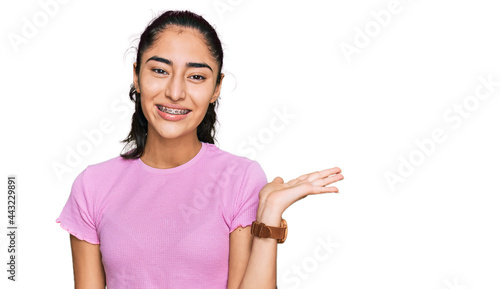 Hispanic teenager girl with dental braces wearing casual clothes smiling cheerful presenting and pointing with palm of hand looking at the camera.