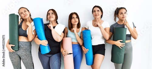 Group of women holding yoga mat standing over isolated background thinking concentrated about doubt with finger on chin and looking up wondering