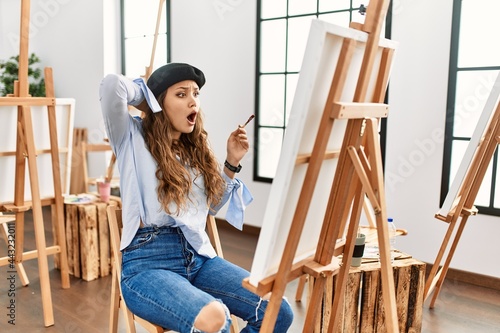 Young hispanic artist woman painting on canvas at art studio crazy and scared with hands on head, afraid and surprised of shock with open mouth