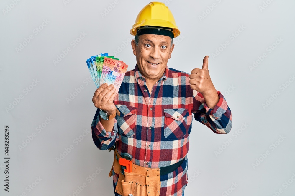 Senior hispanic man wearing handyman uniform and hardhat smiling happy and positive, thumb up doing excellent and approval sign