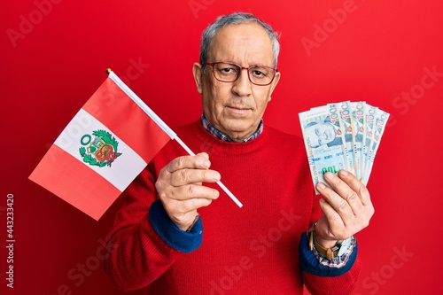 Handsome senior man with grey hair holding peru flag and peruvian sol banknotes relaxed with serious expression on face. simple and natural looking at the camera.