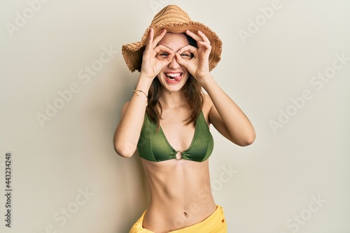 Young brunette woman wearing bikini doing ok gesture like binoculars sticking tongue out, eyes looking through fingers. crazy expression.