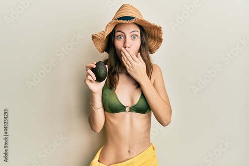 Young brunette woman holding avocado covering mouth with hand  shocked and afraid for mistake. surprised expression