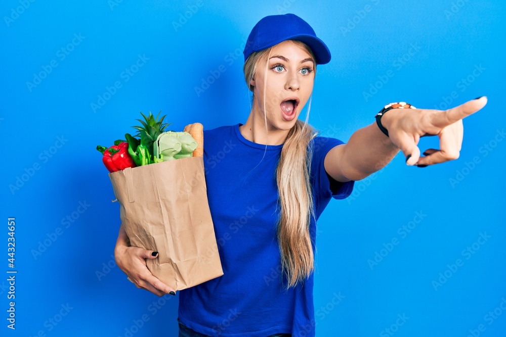 Young caucasian woman wearing courier uniform with groceries from supermarket pointing with finger surprised ahead, open mouth amazed expression, something on the front Young caucasian woman wearing courier uniform with groceries from supermarket pointing with finger surprised ahead, open mouth amazed expression, something on the front