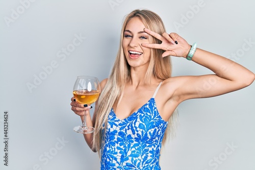 Young beautiful caucasian woman drinking a glass of white wine doing peace symbol with fingers over face, smiling cheerful showing victory