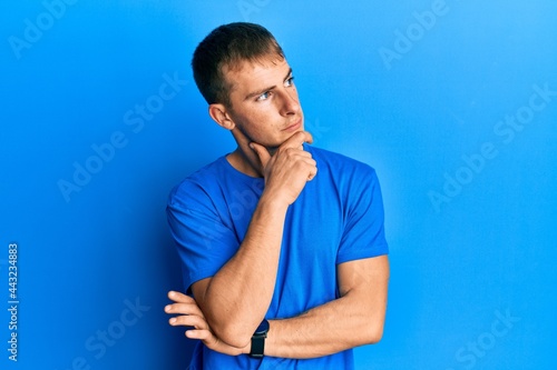 Young caucasian man wearing casual blue t shirt with hand on chin thinking about question, pensive expression. smiling with thoughtful face. doubt concept.