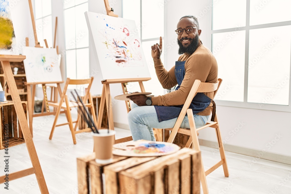 African american artist man painting on canvas at art studio surprised with an idea or question pointing finger with happy face, number one African american artist man painting on canvas at art studio surprised with an idea or question pointing finger with happy face, number one