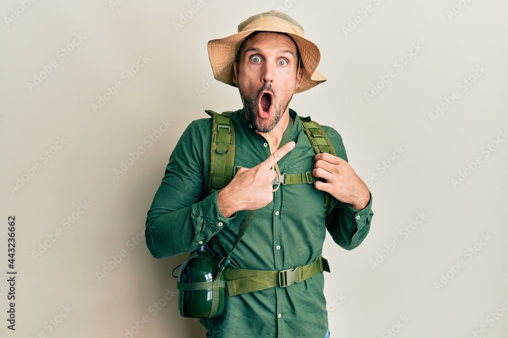 Handsome man with beard wearing explorer hat and backpack surprised pointing with finger to the side, open mouth amazed expression. Handsome man with beard wearing explorer hat and backpack surprised pointing with finger to the side, open mouth amazed expression.