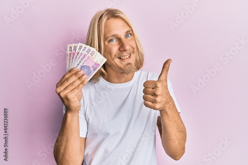 Caucasian young man with long hair holding 20 polish zloty banknotes smiling happy and positive, thumb up doing excellent and approval sign