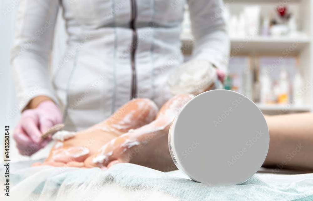 In the foreground is a bank with a white lid without an inscription. On a blurry background, the master applies the cream to the client's heels. Selective focus. In the foreground is a bank with a white lid without an inscription. On a blurry background, the master applies the cream to the client's heels. Selective focus.