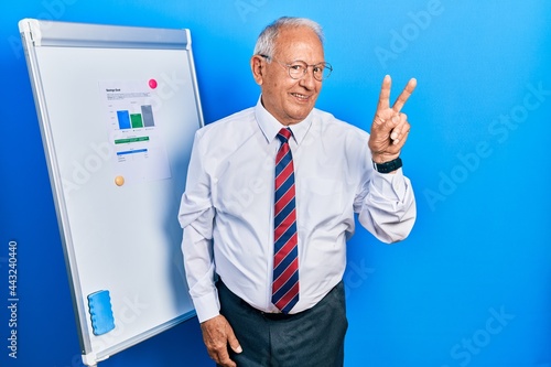 Senior man with grey hair standing by business blackboard smiling looking to the camera showing fingers doing victory sign. number two.