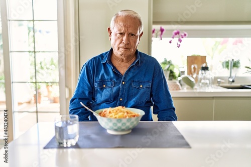 Senior man with grey hair eating pasta spaghetti at home skeptic and nervous, frowning upset because of problem. negative person.
