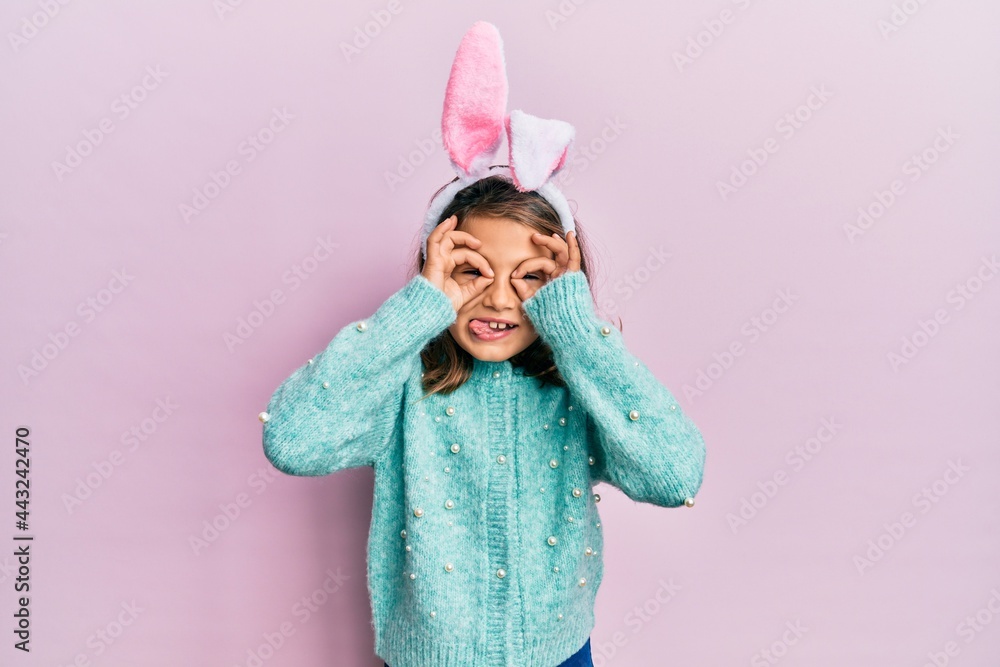 Fotografie Little beautiful girl wearing cute easter bunny ears doing ok gesture like binoculars sticking tongue out, eyes looking through fingers