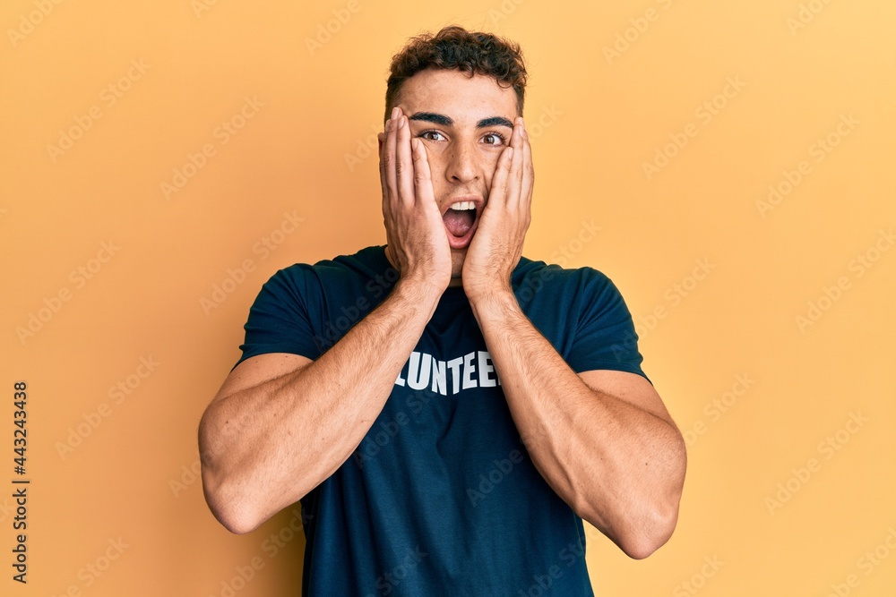 Hispanic young man wearing volunteer t shirt afraid and shocked, surprise and amazed expression with hands on face Hispanic young man wearing volunteer t shirt afraid and shocked, surprise and amazed expression with hands on face