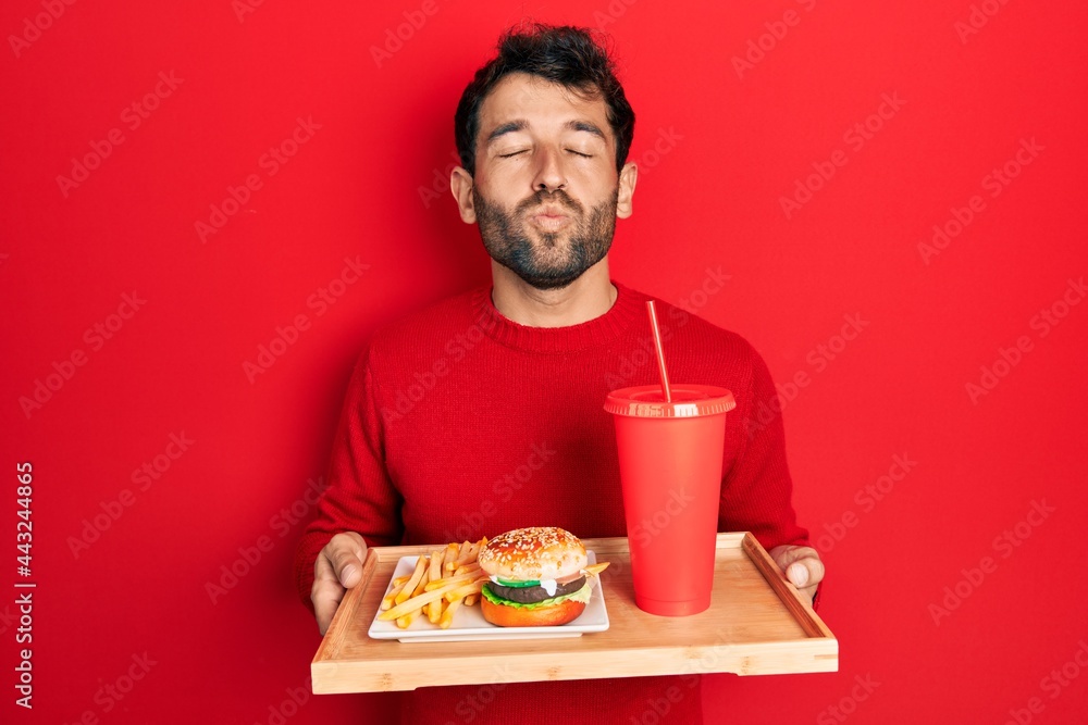 Handsome man with beard eating a tasty classic burger with fries and soda looking at the camera blowing a kiss being lovely and sexy. love expression. Handsome man with beard eating a tasty classic burger with fries and soda looking at the camera blowing a kiss being lovely and sexy. love expression.