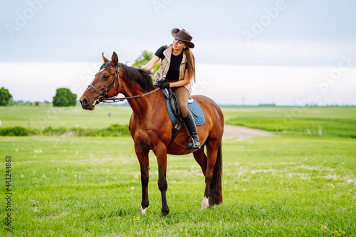 Young woman dressed in riding clothes and hat riding brown horse in green field