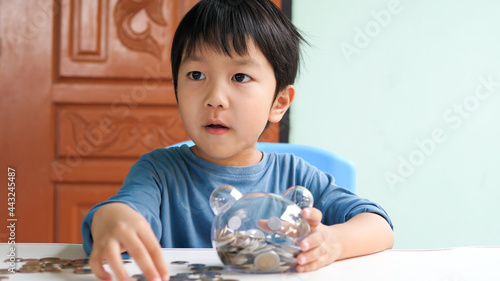 Asian cute child holding transparent piggy bank and looking with surprised face at home. Concept of money saving for future, financial business, investment, banking.