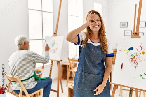 Hispanic woman wearing apron at art studio doing ok gesture with hand smiling, eye looking through fingers with happy face.