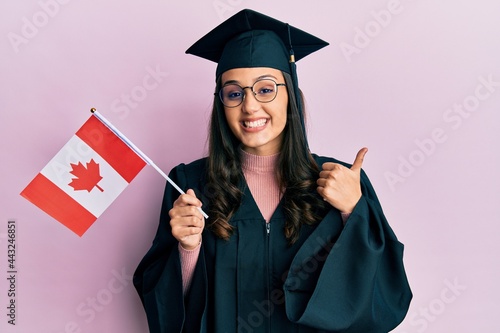 Young hispanic woman wearing graduation uniform holding canada flag smiling happy and positive, thumb up doing excellent and approval sign