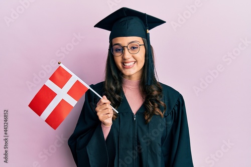 Young hispanic woman wearing graduation uniform holding denmark flag looking positive and happy standing and smiling with a confident smile showing teeth