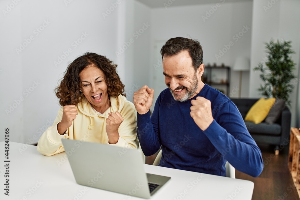 Middle age hispanic couple smiling happy with fists raised up. Sitting on the table using laptop at home. Middle age hispanic couple smiling happy with fists raised up. Sitting on the table using laptop at home.