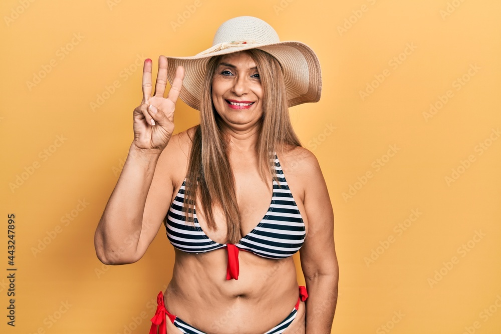 Middle age hispanic woman wearing bikini and summer hat showing and pointing up with fingers number three while smiling confident and happy. Middle age hispanic woman wearing bikini and summer hat showing and pointing up with fingers number three while smiling confident and happy.