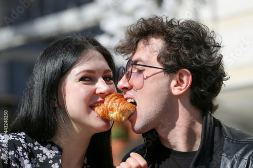 Happy young couple in morning and having breakfast. Cheerful man and woman bite a croissant at the same time.