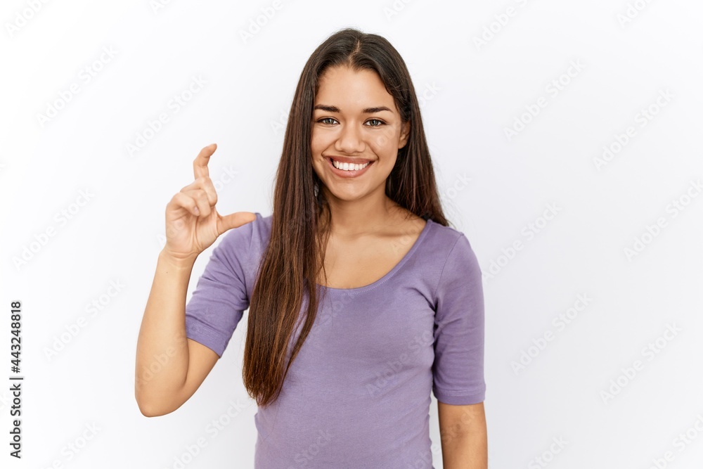 Young brunette woman standing by isolated background smiling and confident gesturing with hand doing small size sign with fingers looking and the camera. measure concept.