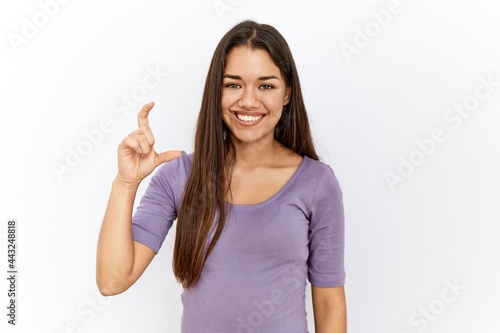 Young brunette woman standing by isolated background smiling and confident gesturing with hand doing small size sign with fingers looking and the camera. measure concept.