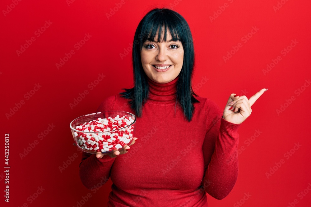 Young hispanic woman holding bowl of pills smiling happy pointing with hand and finger to the side Young hispanic woman holding bowl of pills smiling happy pointing with hand and finger to the side