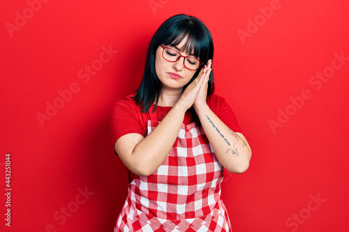 Young hispanic woman wearing cook apron and glasses sleeping tired dreaming and posing with hands together while smiling with closed eyes.
