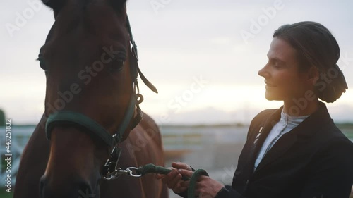Horse Owner with her seal brown horse holding its lead rope. Smiling at her horse and looking at it with love. Evevning time. Horse head closeup view. Horse riding for leisure concept. photo