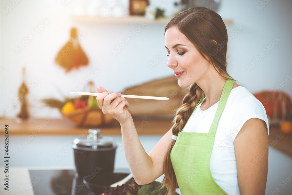 An attractive young woman preparing soup by new keto recipe while standing and smiling in sunny kitchen. Cooking and householding concepts An attractive young woman preparing soup by new keto recipe while standing and smiling in sunny kitchen. Cooking and householding concepts