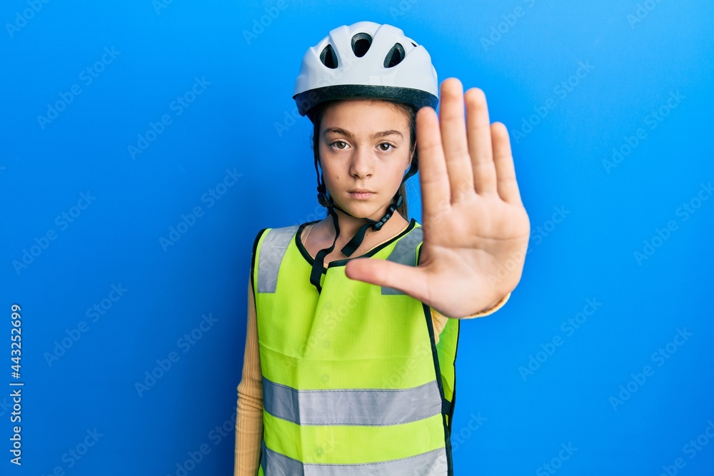 Beautiful brunette little girl wearing bike helmet and reflective vest doing stop sing with palm of the hand. warning expression with negative and serious gesture on the face. Beautiful brunette little girl wearing bike helmet and reflective vest doing stop sing with palm of the hand. warning expression with negative and serious gesture on the face.
