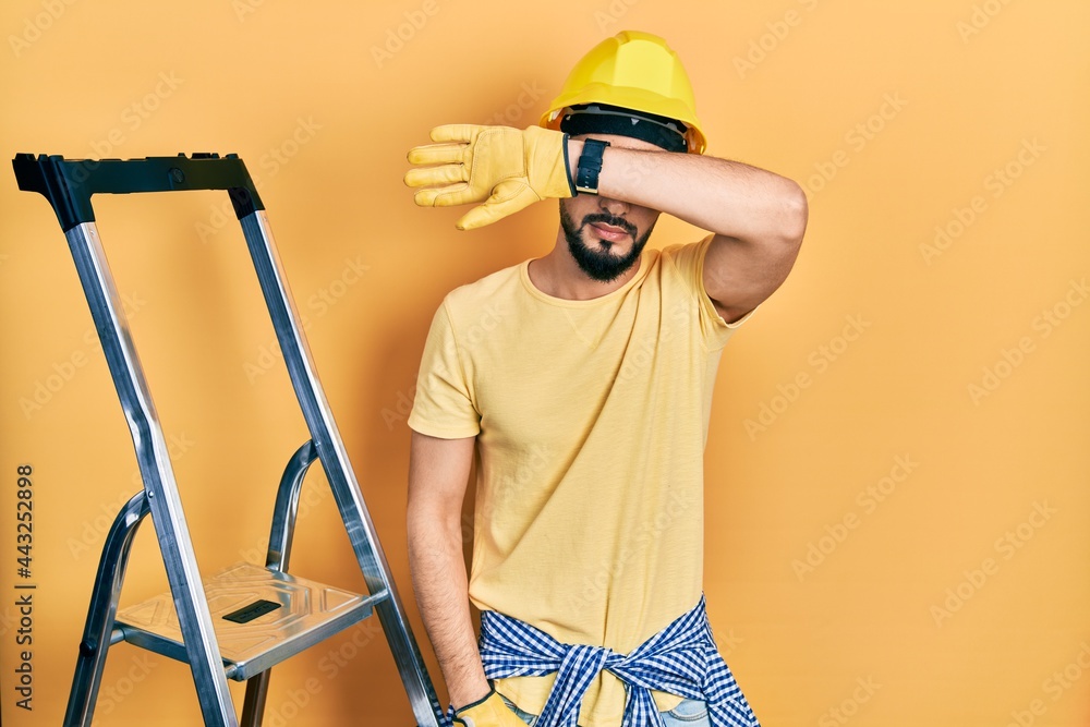 Handsome man with beard by construction stairs wearing hardhat covering eyes with arm, looking serious and sad. sightless, hiding and rejection concept Handsome man with beard by construction stairs wearing hardhat covering eyes with arm, looking serious and sad. sightless, hiding and rejection concept