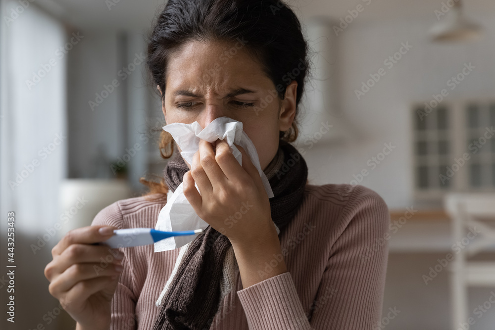 Unhealthy young latin woman wiping runny nose with paper tissue, measuring body temperature with electronic device. Worrying millennial hispanic lady having first flue grippe corona virus symptoms.