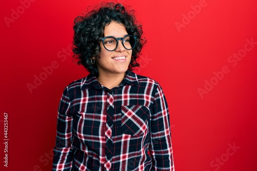 Young hispanic woman with curly hair wearing casual clothes and glasses looking away to side with smile on face, natural expression. laughing confident.