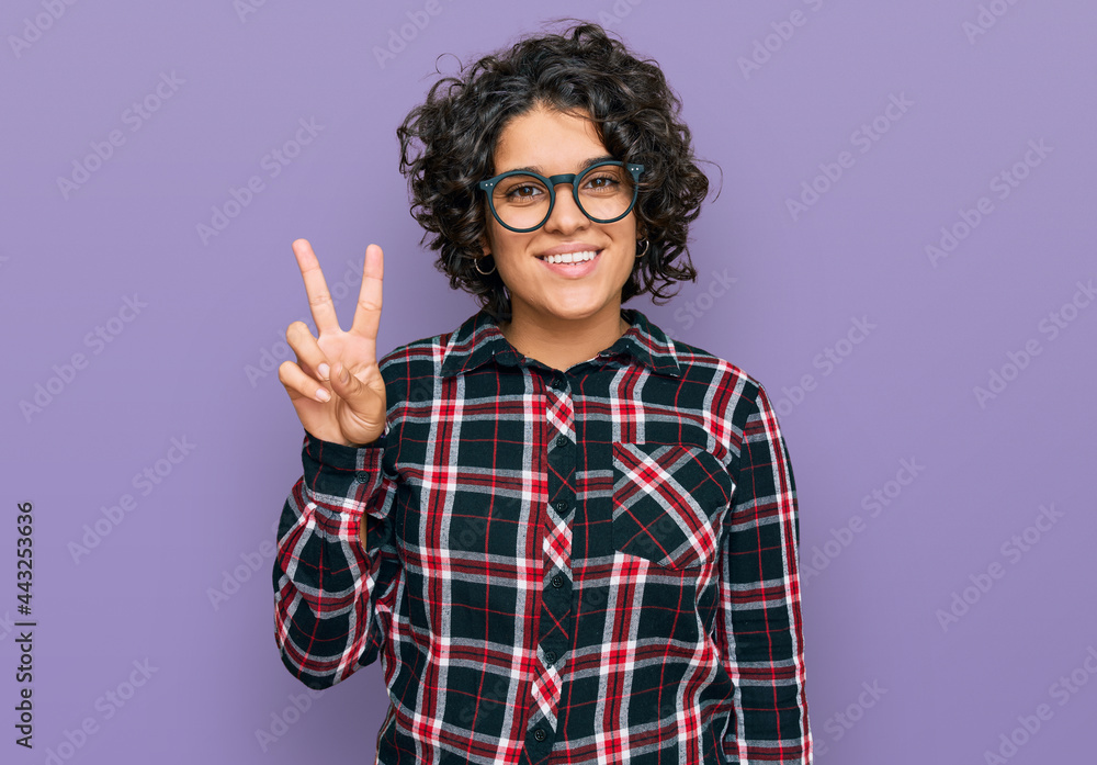 Young hispanic woman with curly hair wearing casual clothes and glasses smiling with happy face winking at the camera doing victory sign. number two. Young hispanic woman with curly hair wearing casual clothes and glasses smiling with happy face winking at the camera doing victory sign. number two.