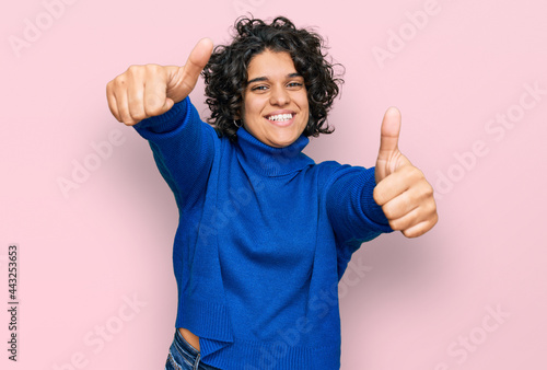 Young hispanic woman with curly hair wearing turtleneck sweater approving doing positive gesture with hand  thumbs up smiling and happy for success. winner gesture.