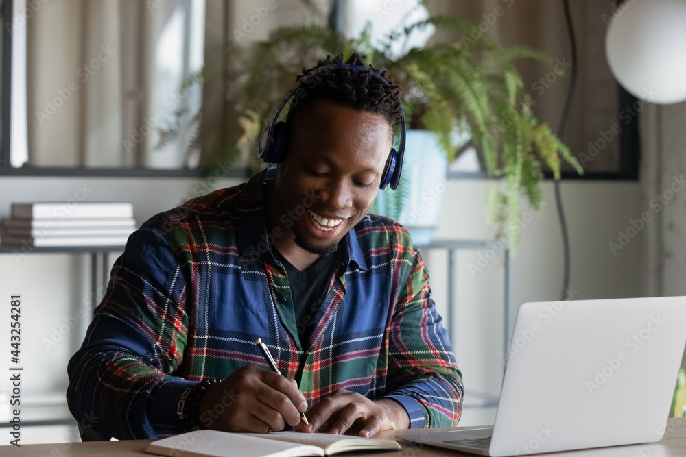 Smiling African American man in headphones taking notes, studying online, intern watching webinar in office using laptop, happy motivated student engaged in internet course conference seminar Smiling African American man in headphones taking notes, studying online, intern watching webinar in office using laptop, happy motivated student engaged in internet course conference seminar