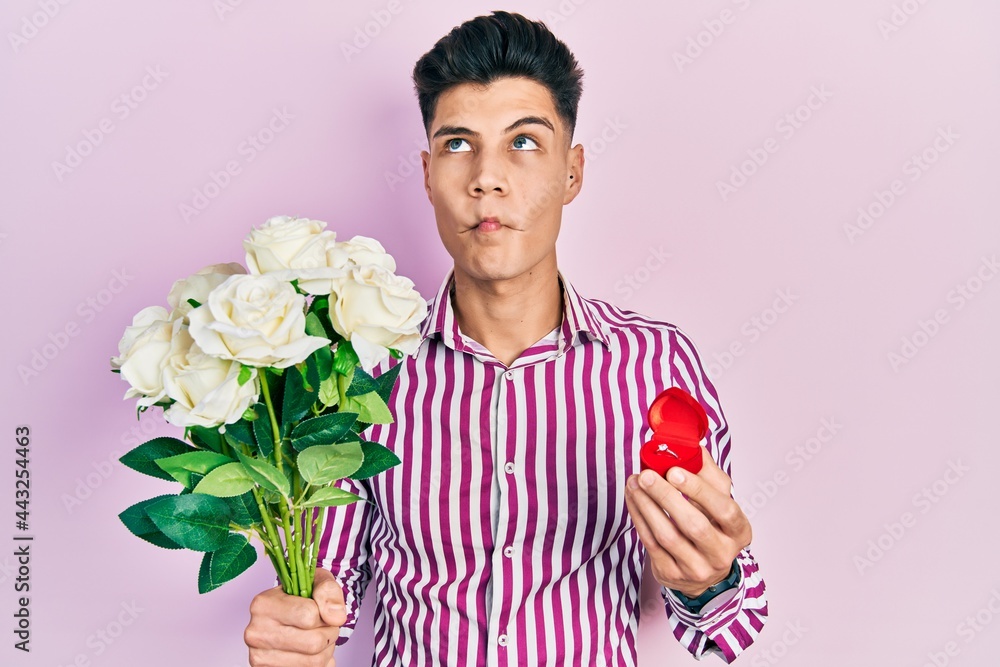 Young hispanic man holding bouquet of flowers and wedding ring making fish face with mouth and squinting eyes, crazy and comical. Young hispanic man holding bouquet of flowers and wedding ring making fish face with mouth and squinting eyes, crazy and comical.