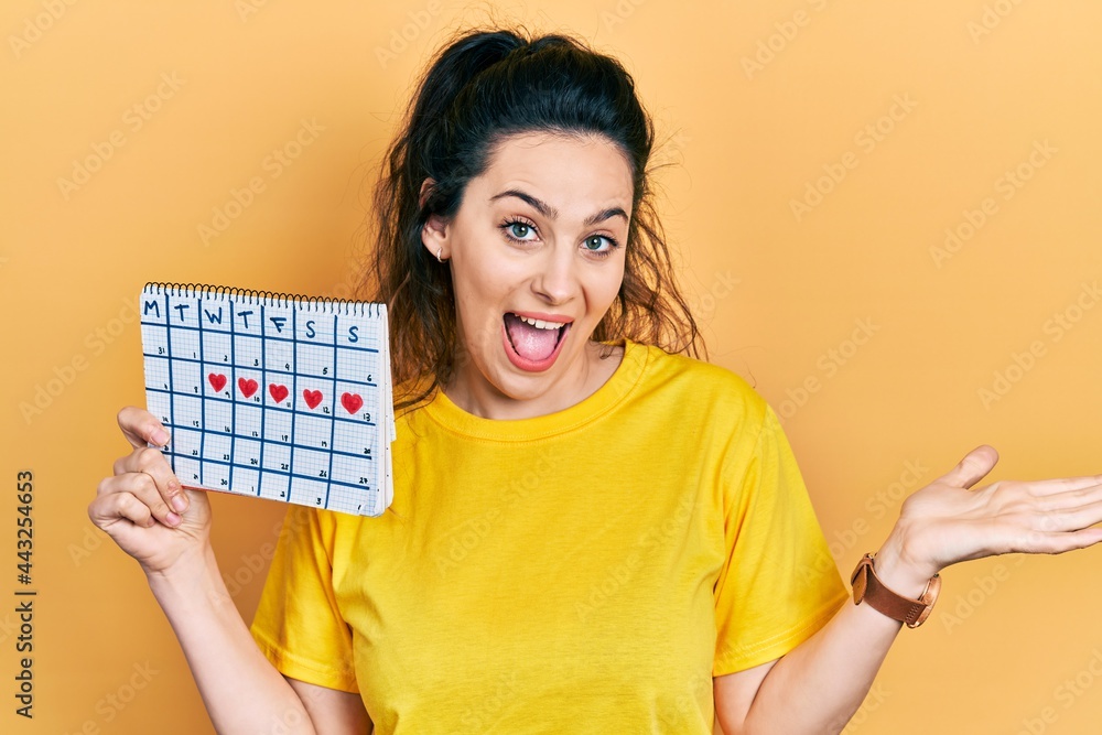 Young hispanic woman holding heart calendar celebrating achievement with happy smile and winner expression with raised hand Young hispanic woman holding heart calendar celebrating achievement with happy smile and winner expression with raised hand