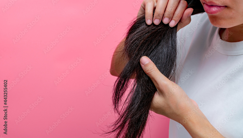 Fototapeta premium Asian woman with dry lips holding damaged hair on pink background. Dry and brittle black long hair needs shampoo and conditioner for spa treatment. Hair loss and thin hair problems in women.