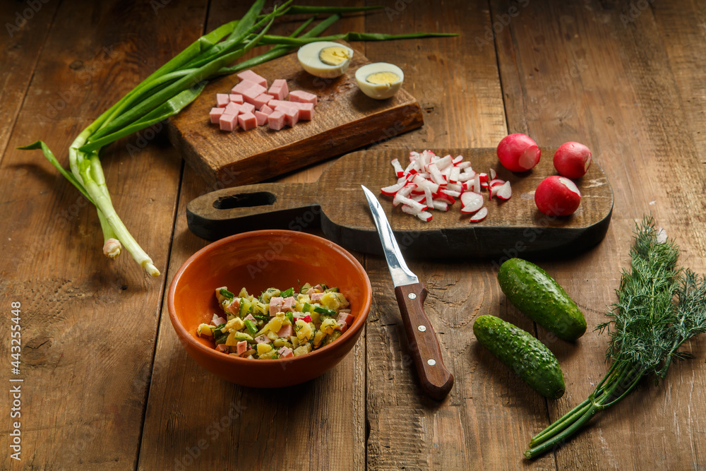 Okroshka in a clay plate on a table on a wooden table next to vegetables on boards and a knife.