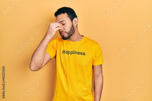 Hispanic man with beard wearing t shirt with happiness word message tired rubbing nose and eyes feeling fatigue and headache. stress and frustration concept.