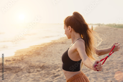 Young sporty woman doing fitness with stretch ropes at sunset. Beach shoulders training with elastic rubber bands. Healthy active lifestyle concept