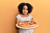 Young little girl with afro hair holding italian pizza clueless and confused expression. doubt concept.