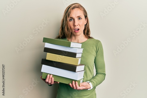 Young blonde woman holding a pile of books in shock face, looking skeptical and sarcastic, surprised with open mouth